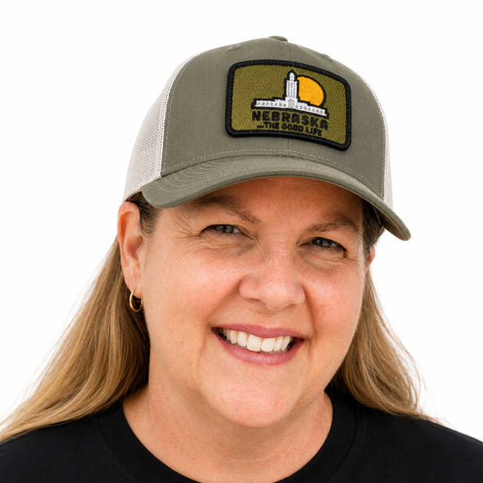 A smiling woman wearing an olive green trucker hat with a rectangular patch that reads 'NEBRASKA THE GOOD LIFE' and shows her face from the chest up against a plain white background.