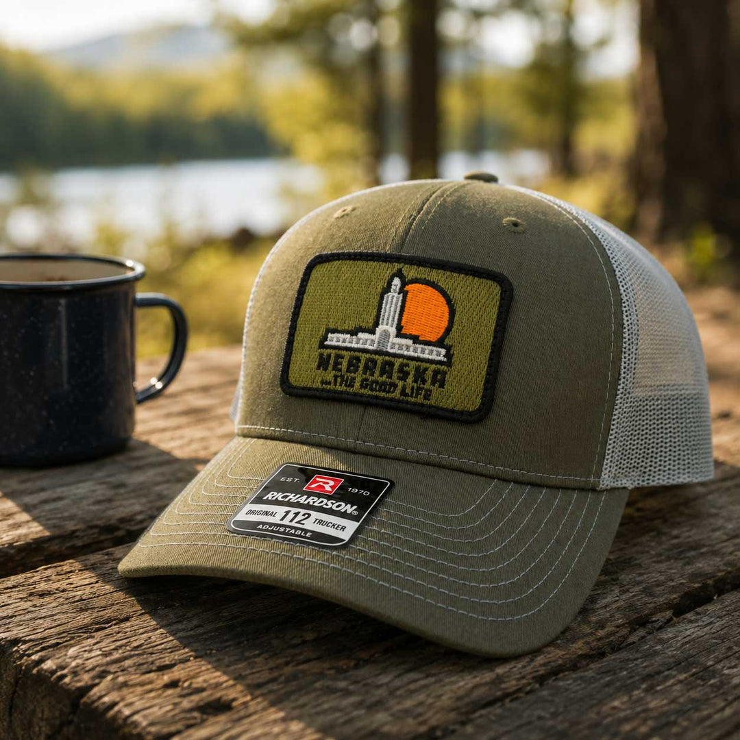 An olive green Richardson 112 trucker hat with a Nebraska patch, resting on a rustic wooden table next to a black mug with a blurred outdoor lake and forest background.