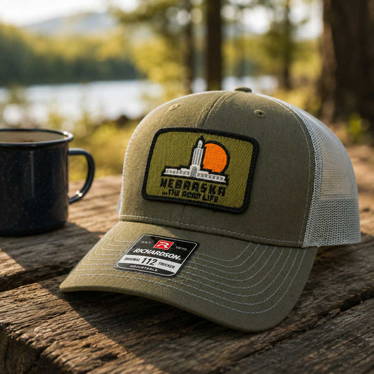 An olive green Richardson 112 trucker hat with a Nebraska patch, resting on a rustic wooden table next to a black mug with a blurred outdoor lake and forest background.