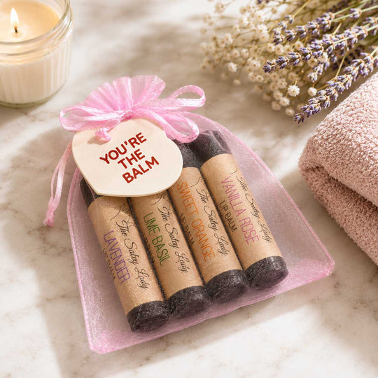 Set of lip balms in a pink bag with a tag, surrounded by lavender and a candle on a marble surface.