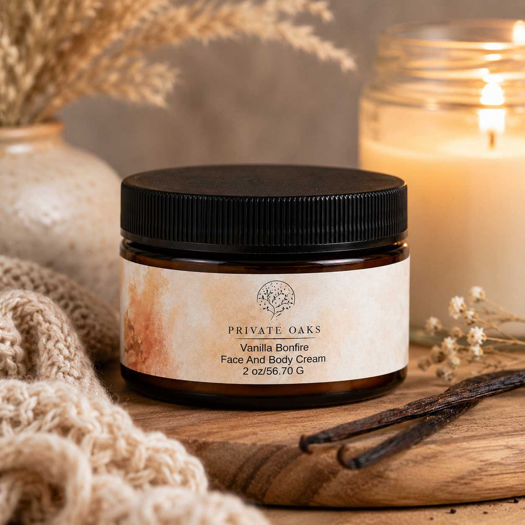 A jar of Private Oaks Vanilla Bonfire Face And Body Cream sits on a wooden surface surrounded by cozy props, including a knitted beige scarf, dried flowers, vanilla beans, and a lit candle in the background.