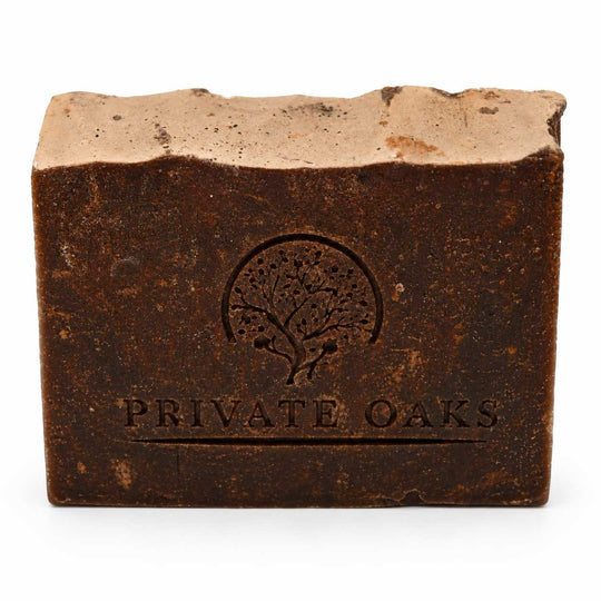 A rectangular bar of soap with a rustic, textured dark brown finish and a lighter beige top surface. The front is embossed with a tree logo and the brand name 'PRIVATE OAKS' in a serif font, set against a plain white background.