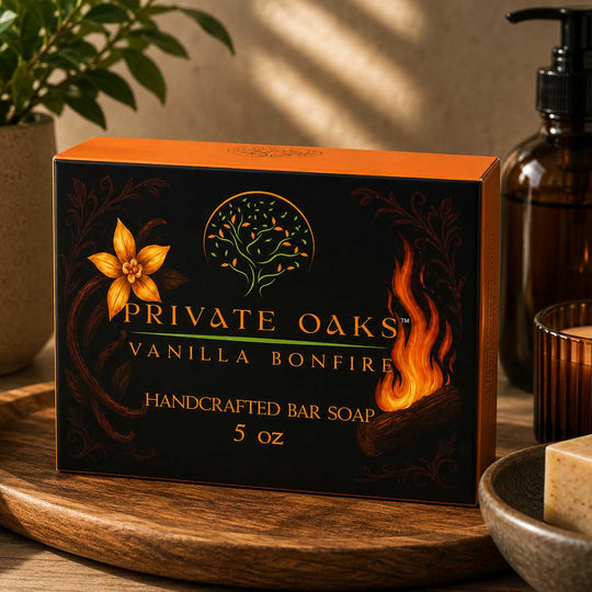 A boxed bar of soap labeled Private Oaks Vanilla Bonfire sits on a wooden tray in a styled setting with a plant, amber bottle, candle, and bowl nearby.