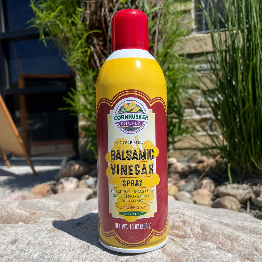 A 10 oz can of Cornhusker Kitchen Gourmet Balsamic Vinegar Spray standing on a stone surface outdoors with plants and a house in the background.