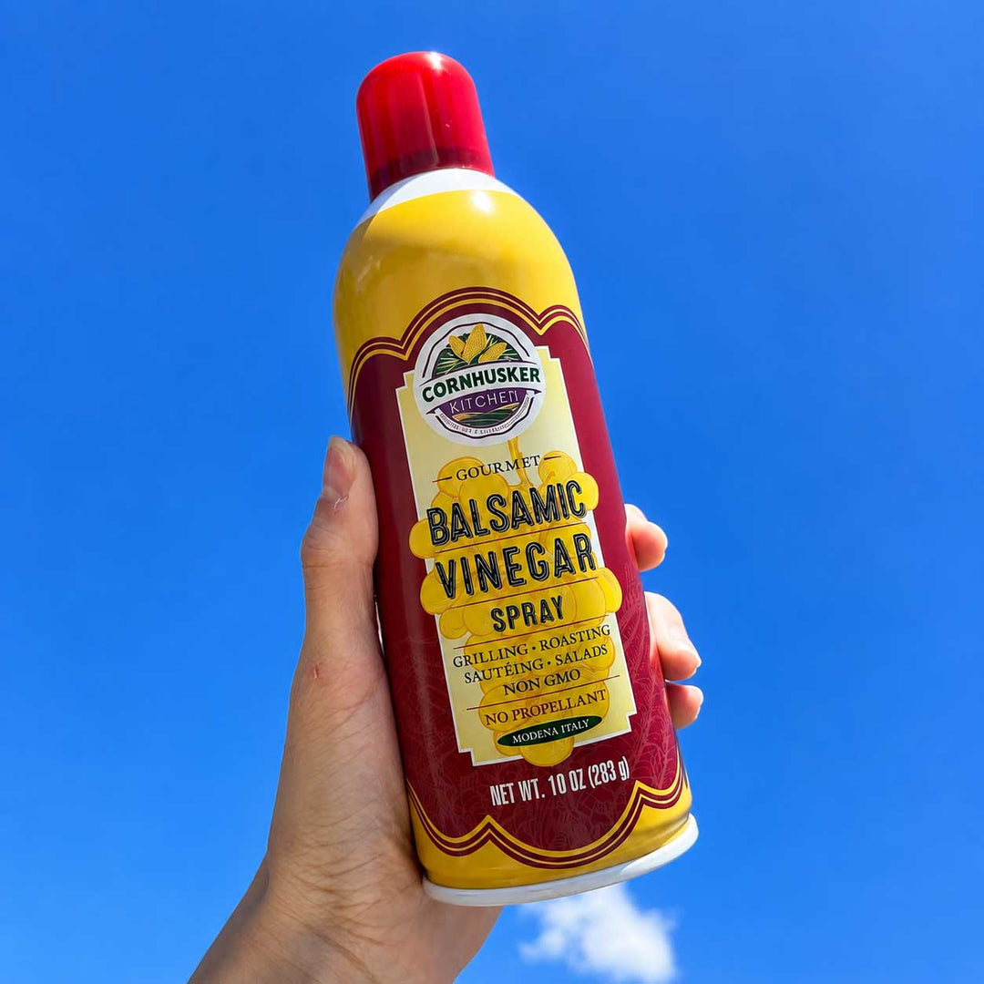 A hand holding a can of Cornhusker Gourmet Balsamic Vinegar Spray against a bright blue sky background.