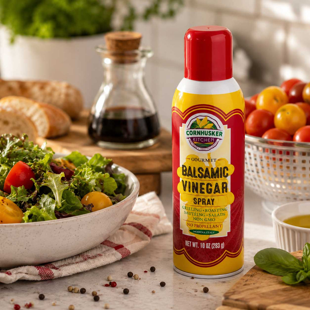 A bottle of Cornusker Vinegar Gourmet Balsamic Vinegar Spray stands on a kitchen counter beside a salad bowl, bread, a glass bottle of dark liquid, and tomatoes. The product bottle is yellow and red with a white label and a red cap.