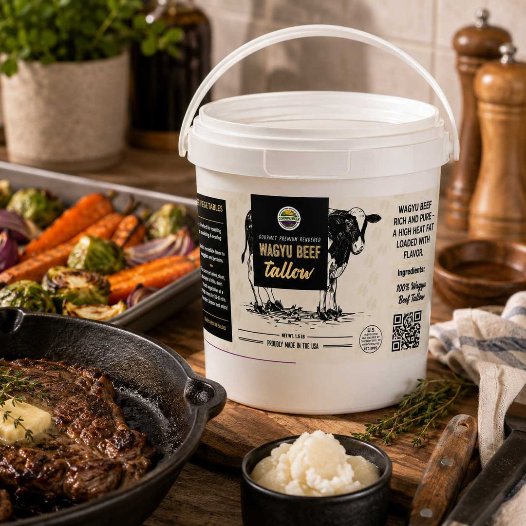 A white plastic tub of Wagyu beef fat by Wagyu Beef Co. sits on a rustic wooden table surrounded by cooked steak, mashed potatoes, carrots, and herbs, with wooden pepper mills in the background.
