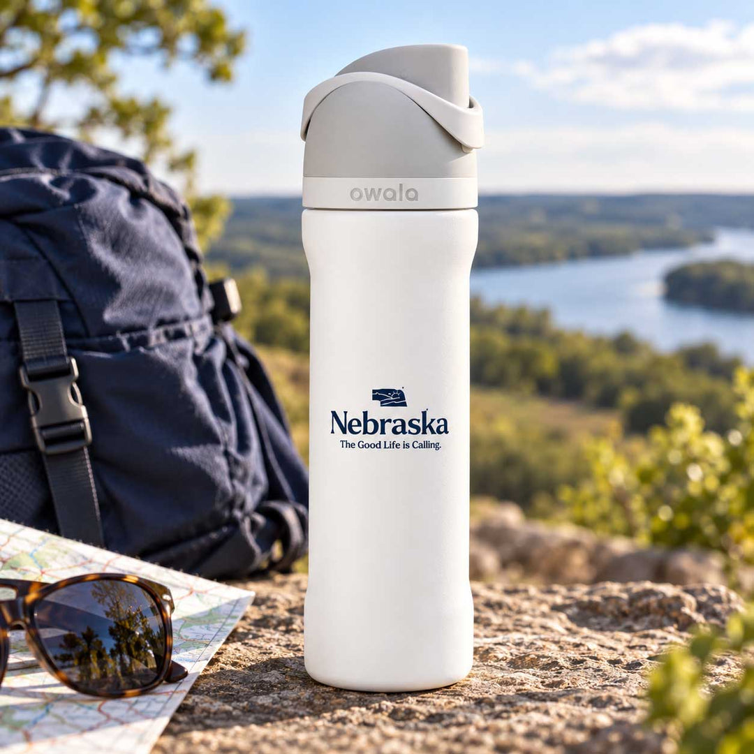 A white Owala insulated water bottle with a gray lid stands on a rocky outdoor overlook next to a navy backpack, sunglasses, and a map, with a river and forest in the background. The bottle is printed with the Nebraska logo.