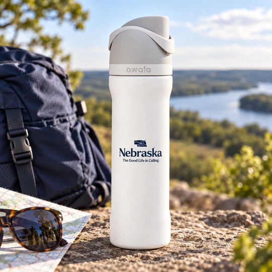 A white Owala insulated water bottle with a gray lid stands on a rocky outdoor overlook next to a navy backpack, sunglasses, and a map, with a river and forest in the background. The bottle is printed with the Nebraska logo.