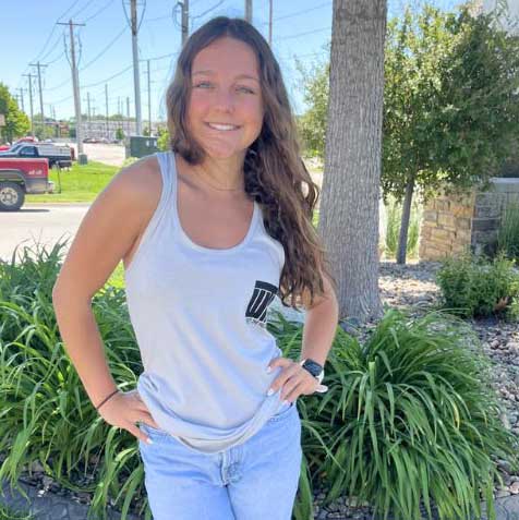 A woman smiling outdoors wearing a light gray tank top with a black graphic logo on the left chest and light blue jeans, standing in front of green plants and trees.