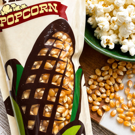 Popcorn bag with corn kernels, a bowl of popcorn, and scattered kernels on a wooden surface.