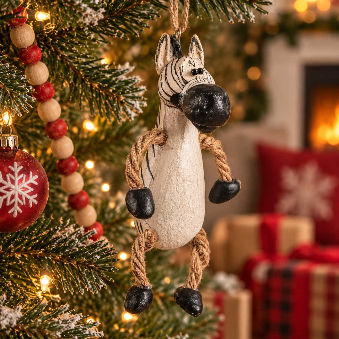 A zebra-shaped hanging ornament made from a cream-colored wooden block with black painted stripes, a black nose, and rope limbs, hanging on a decorated Christmas tree with warm lights and holiday decor in the background.