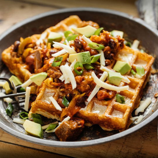 Plate of freshly made waffles topped with avocado, cheese, beef tips, and green onions. 